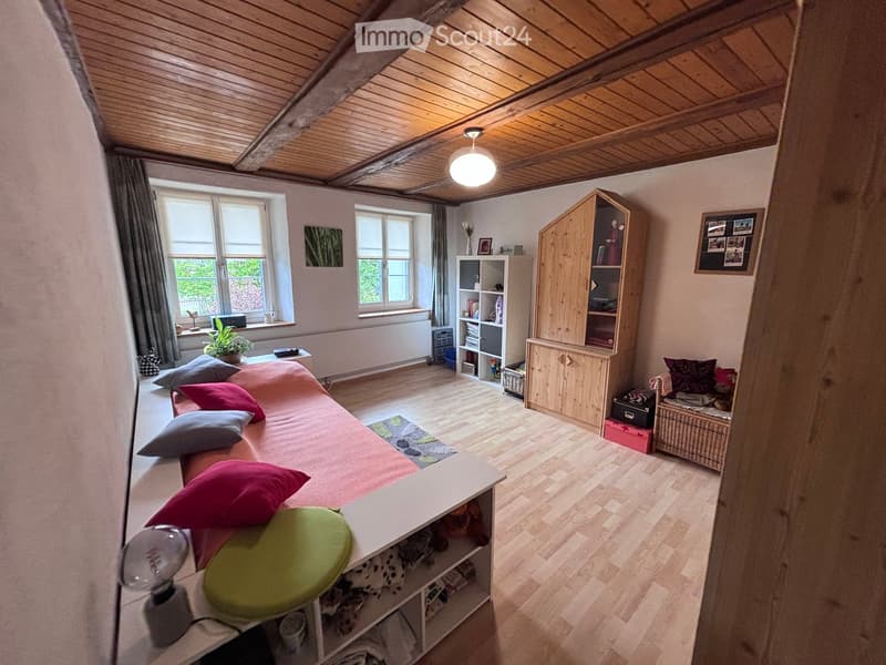 Cozy bedroom with wooden floors, slanted wooden ceiling, large windows, a sofa with colorful pillows, a shelf, a wooden cabinet, a small plant, and a picture frame on the wall.