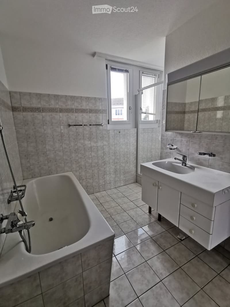 Bathroom with a bathtub, sink, mirror, and tiled walls and floor. Windows with blinds provide natural light.