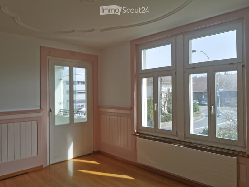 Empty room with wooden flooring, large windows, and a door leading to a balcony. The walls are painted in a light pink color with decorative molding. Natural light streams in from the windows, illuminating the space.