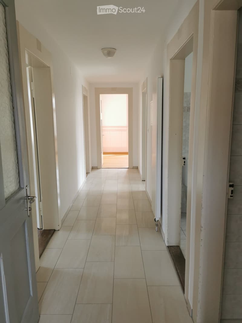 A long, narrow hallway with tiled flooring and white walls, featuring several doors and a visible light fixture on the ceiling.