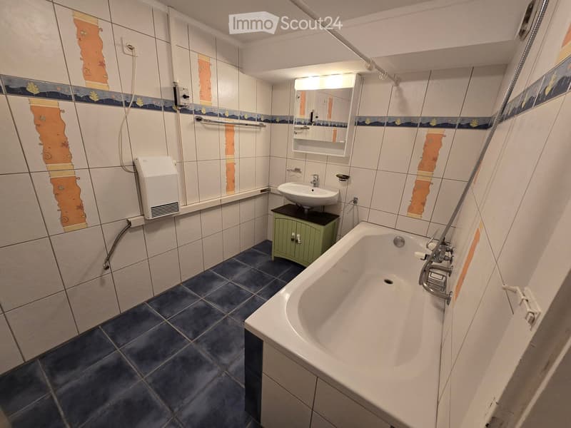 Bathroom with white tiled walls, a bathtub, a sink with a small green cabinet underneath, a mirror, and a handrail for accessibility.