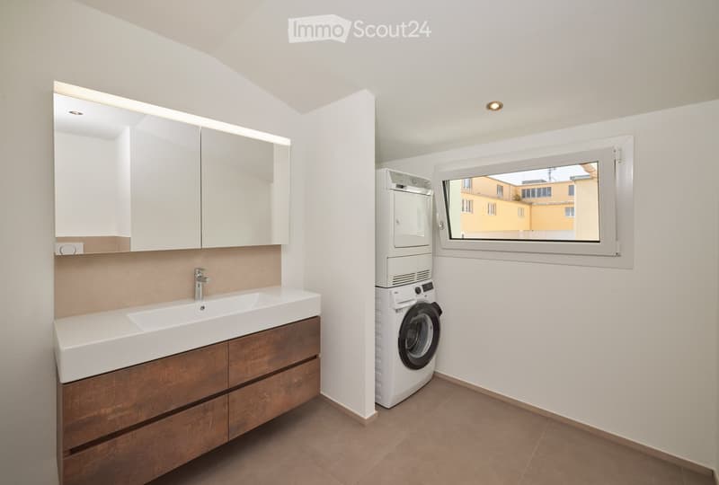 Modern bathroom with a large sink, wooden cabinet, mirror, and a private washing machine. The room features a window with a view of a building outside.