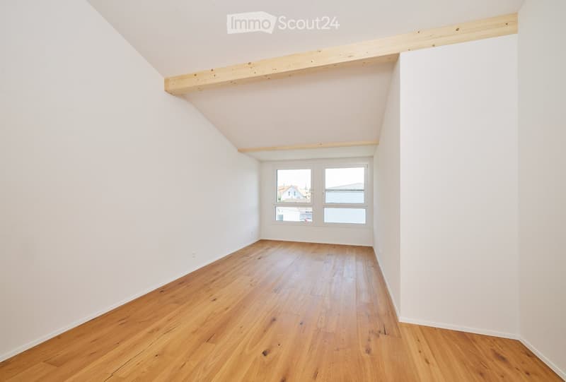 Empty room with wooden flooring and white walls, featuring a slanted ceiling and a window with a view of the outside.