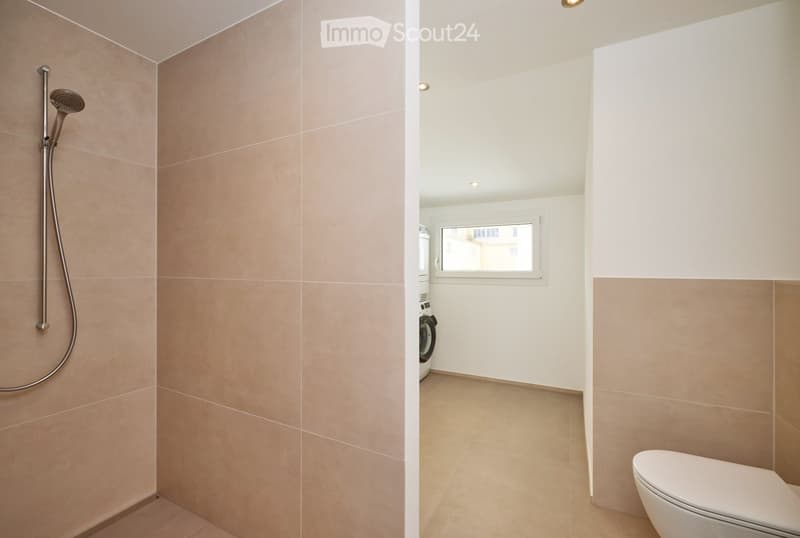 Modern bathroom with beige tiled walls, a shower area, a toilet, and a washing machine visible in the background.