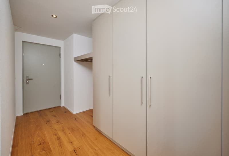 A narrow hallway with light wooden flooring, white walls, built-in white cabinets with silver handles, and a closed door at the end.