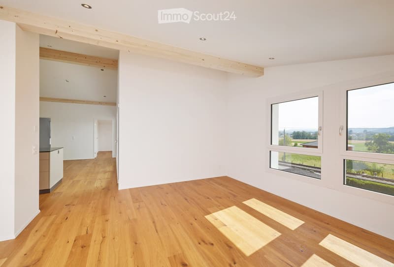 Empty room with light wooden flooring, white walls, and large windows offering a scenic view. The space is unfurnished, with visible structural beams and recessed lighting.
