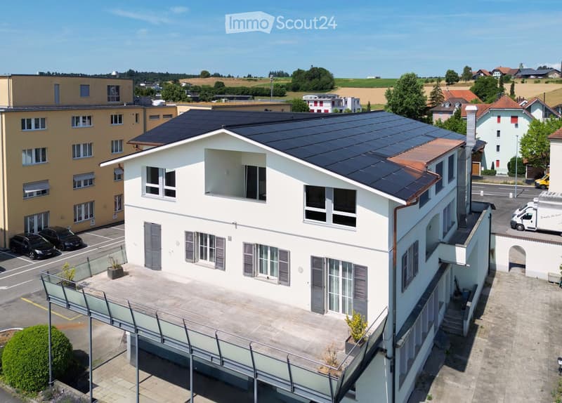 A modern, multi-story building with a flat roof covered in solar panels, surrounded by other structures and greenery.