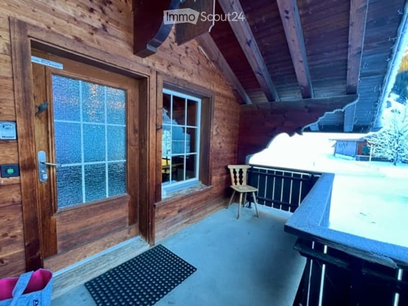 Wooden balcony with a door leading inside, a small chair, and a doormat. Snow-covered ground visible in the background.