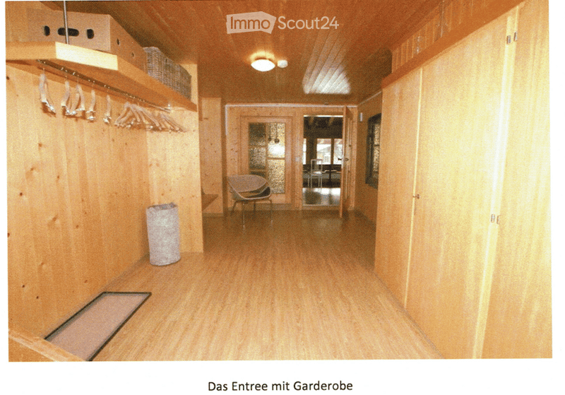 A narrow hallway with wooden walls and flooring, featuring built-in coat racks, a small chair, and a trash bin. The hallway leads to an open doorway with glass panels.