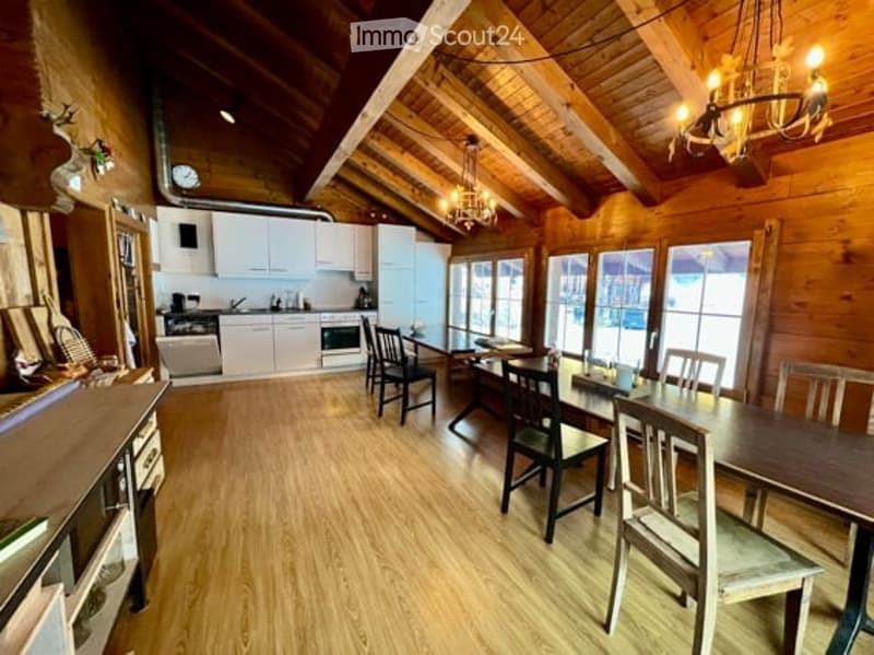 Bright, open-plan living and dining area with exposed wooden beams, white kitchen cabinets, a dining table with chairs, and a chandelier hanging from the ceiling.