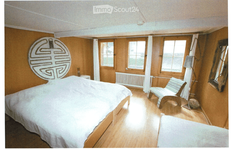 Cozy bedroom with wooden walls and floor, large bed with white bedding, a wooden chair, a nightstand, and a radiator under the windows.