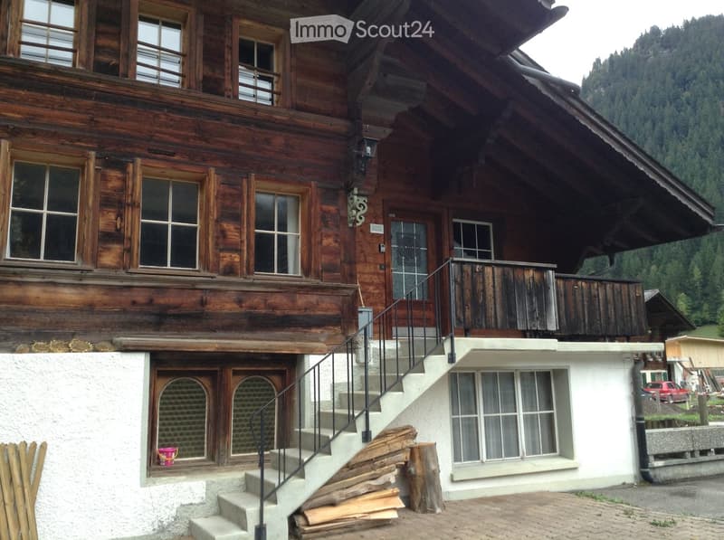A two-story wooden house with a steep roof, multiple windows, and a staircase leading to an upper balcony. The lower level has a white exterior with a small window and a door.