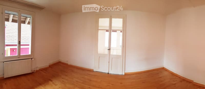 Empty room with wooden flooring, white walls, and large windows offering a view of the roof and surrounding buildings.