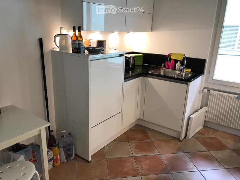 Modern kitchen with white cabinets, black countertops, and a stainless steel sink. Visible items include a refrigerator, a kettle, bottles, and a small table with a stool.