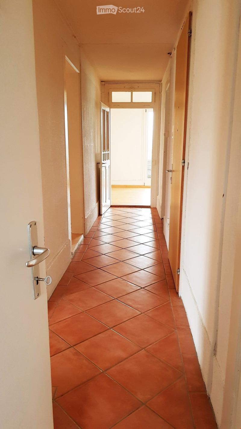 A narrow hallway with tiled flooring and beige walls, featuring several doors and a window at the end.