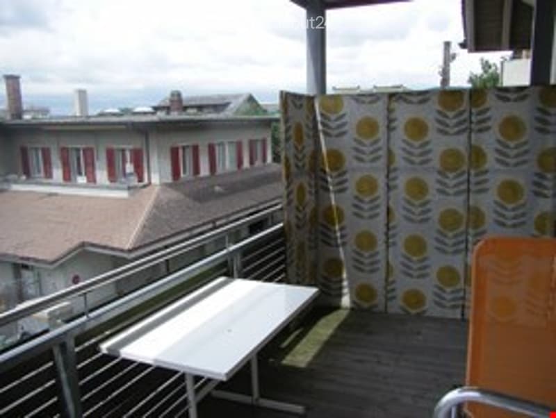 Balcony with metal railing, white table, orange chair, and a large floral curtain partially covering the view of neighboring buildings.