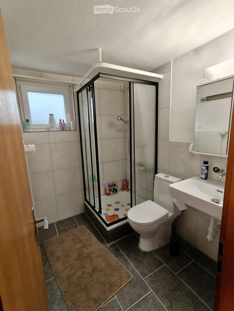 Modern bathroom with a glass-enclosed shower, toilet, sink, and mirror. Includes a small window, a bathroom rug, and various toiletries.