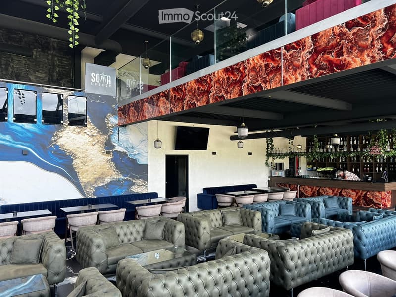 Modern lounge area with grey tufted sofas, tables, chairs, and a bar counter with bottles. Features a television and decorative wall art.