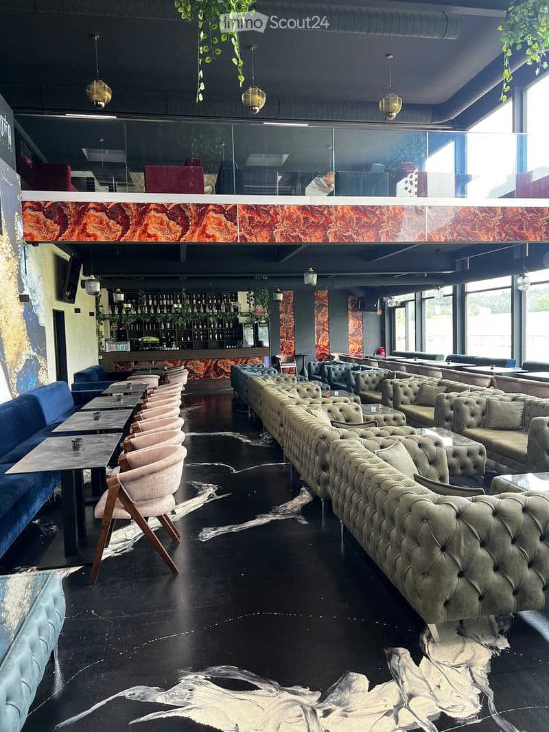 Modern lounge area with dark flooring, large sectional sofa, multiple chairs, tables, and a bar area with shelves of bottles.