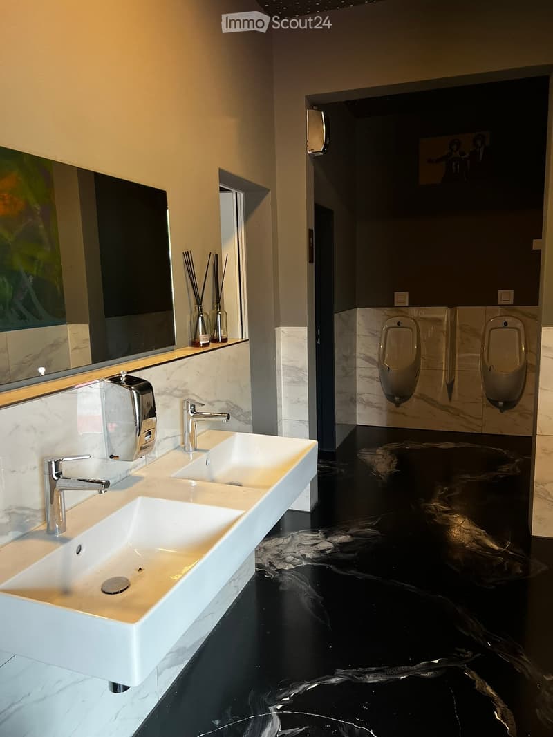 Modern bathroom with white marble sinks, chrome faucets, urinals, and a large mirror. The floor is black with a marble-like pattern.