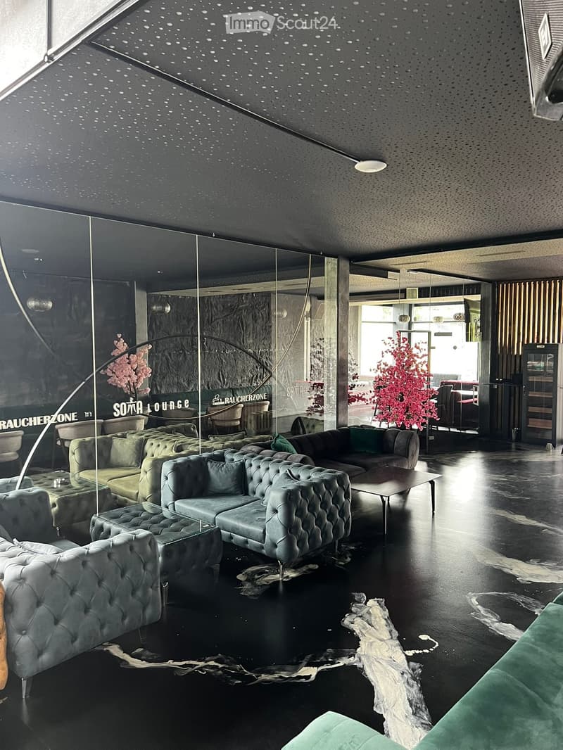 Modern lounge area with dark grey sofas, coffee tables, armchairs, and a sleek floor lamp. The space features a dark polished floor and large glass windows.