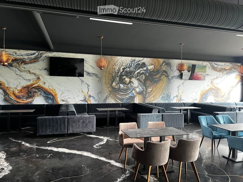 Modern restaurant interior with dark grey sofas, tables, chairs, and a large television screen on the wall.