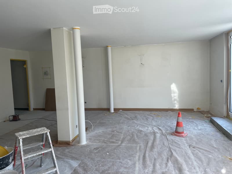 Empty room with white walls, concrete floor, and two large white columns. A small wooden ladder, a traffic cone, and a bucket are visible.