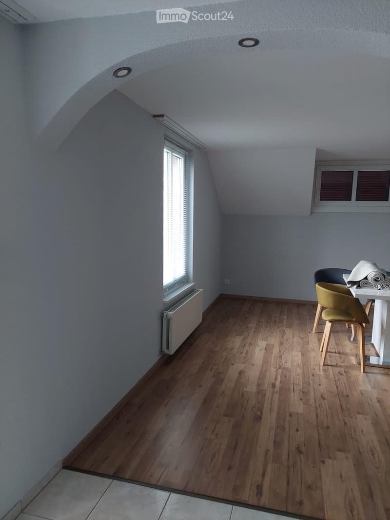 Bright attic room with wooden flooring, slanted ceiling, and two windows. Contains a small table and two chairs.