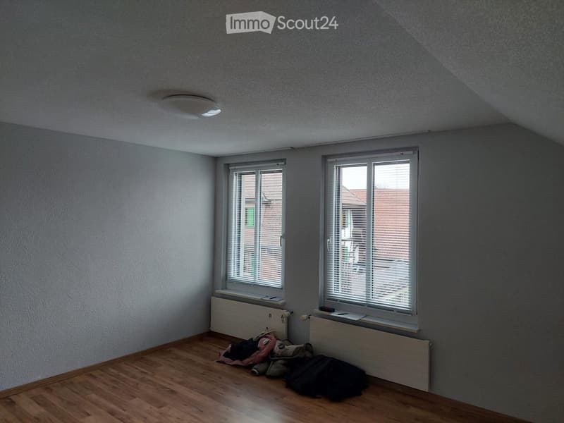 Empty room with wooden flooring, two windows with blinds, radiator, and some scattered items on the floor.