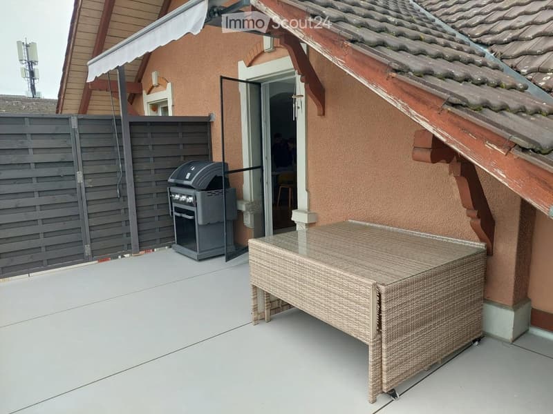 Outdoor patio with a wicker table and a grill, adjacent to a house with a covered entrance.