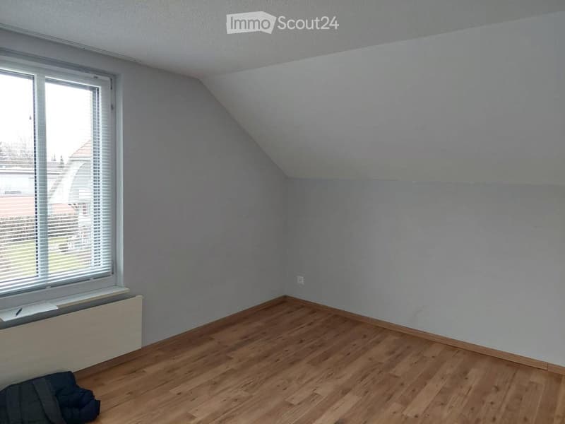 Empty attic room with wooden flooring, slanted ceiling, and a single window with blinds. There is a radiator below the window and a small pile of items on the floor.