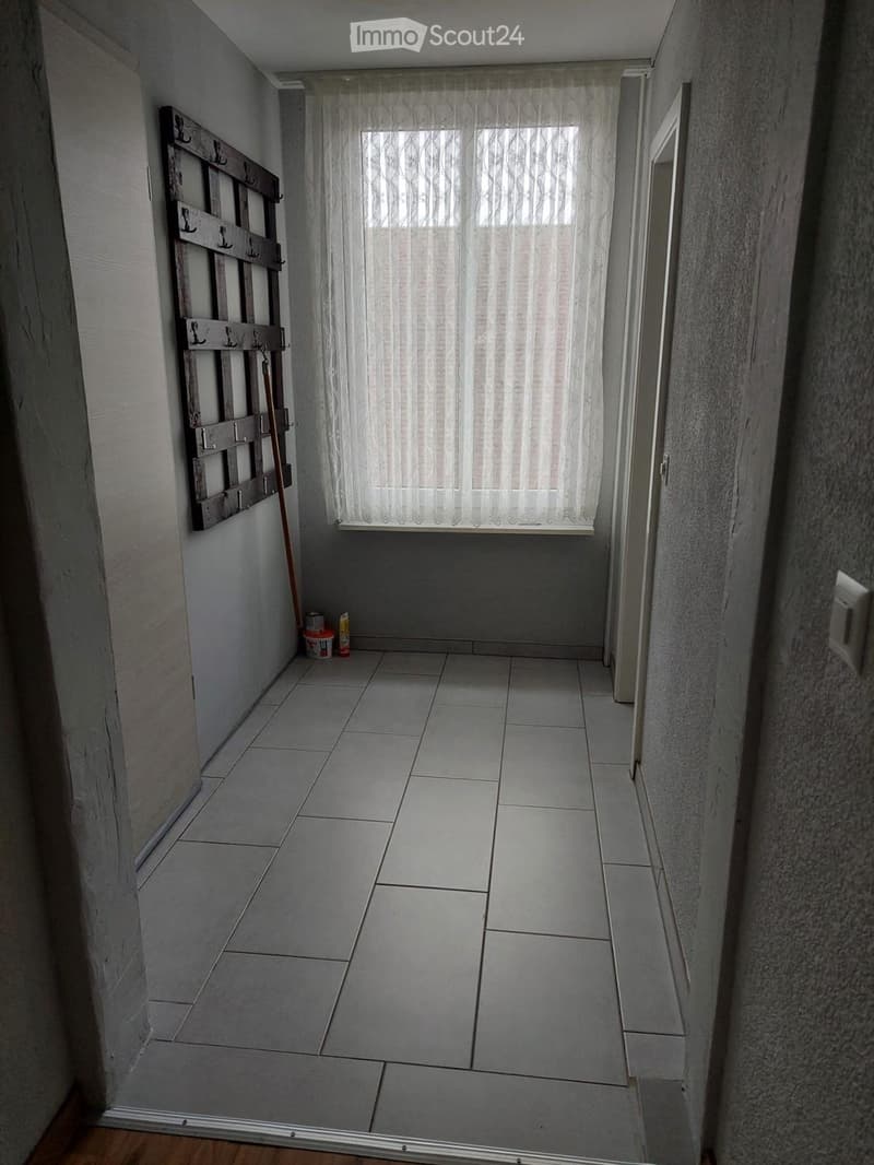 Empty hallway with tiled floor, white walls, and a window with sheer curtains. A coat rack and cleaning supplies are visible on the left side.