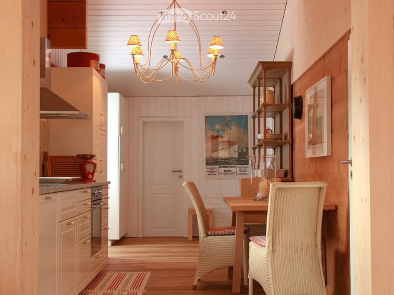 Furnished Room. Cozy kitchen and dining area with light wooden cabinets, a small dining table with two wicker chairs, a chandelier, and a poster on the wall.