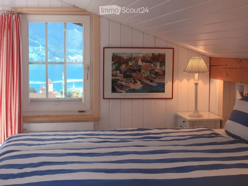 Bedroom. Cozy bedroom with a blue and white striped bedspread, a nightstand with a lamp, a framed picture on the wall, and a window offering a scenic view.