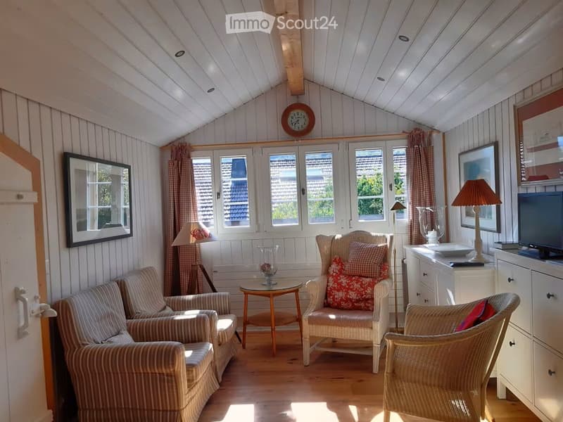 Furnished Room. Cozy living room with wooden floors, white paneled walls, large windows, and a slanted ceiling. Features include a sofa, armchairs, a coffee table, lamps, a television, and decorative items.