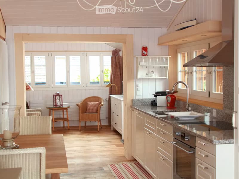Furnished Room. The image shows a modern, well-lit kitchen and dining area with light wood cabinetry, granite countertops, and stainless steel appliances. The kitchen features a sink, faucet, and built-in oven. The dining area includes a wooden table with