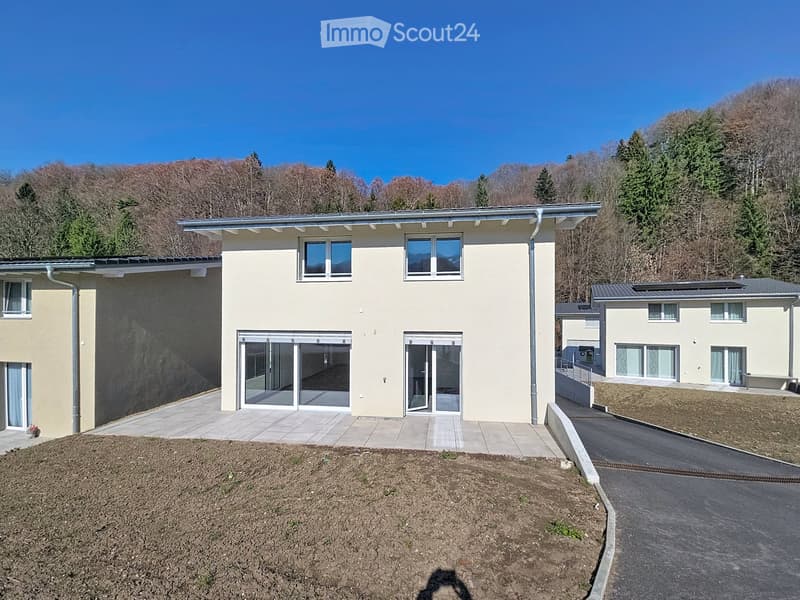 Modern two-story house with light yellow facade, large windows, and sliding glass doors leading to a paved patio area. The house is situated on a raised ground floor with a driveway and surrounded by trees and hills.