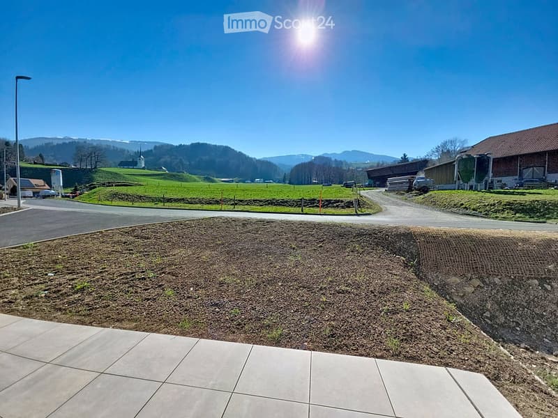 A landscaped outdoor area with a paved walkway, grassy slope, and distant rural buildings and mountains under a clear blue sky.