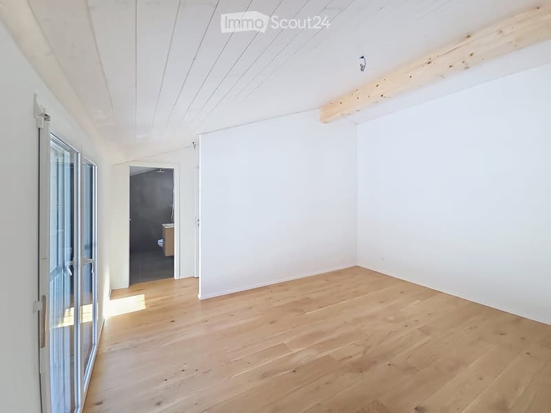 Bright, empty room with light wooden flooring and white walls, featuring a large window and a doorway leading to a small bathroom.