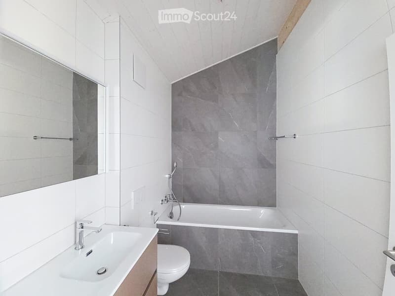 Modern bathroom with white tiles, a large mirror, a sink, a toilet, and a bathtub with a grey accent wall.