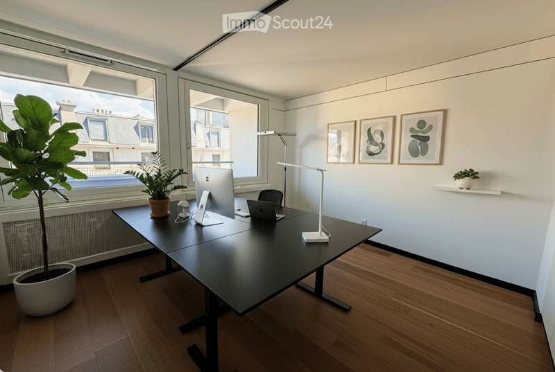 Modern office space with a large black table, laptop, monitor, chair, lamp, potted plants, and framed artwork on the wall.