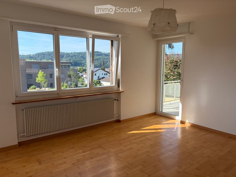 Empty room with wooden flooring, large windows, radiator, and a sliding door leading to a balcony with a scenic view.