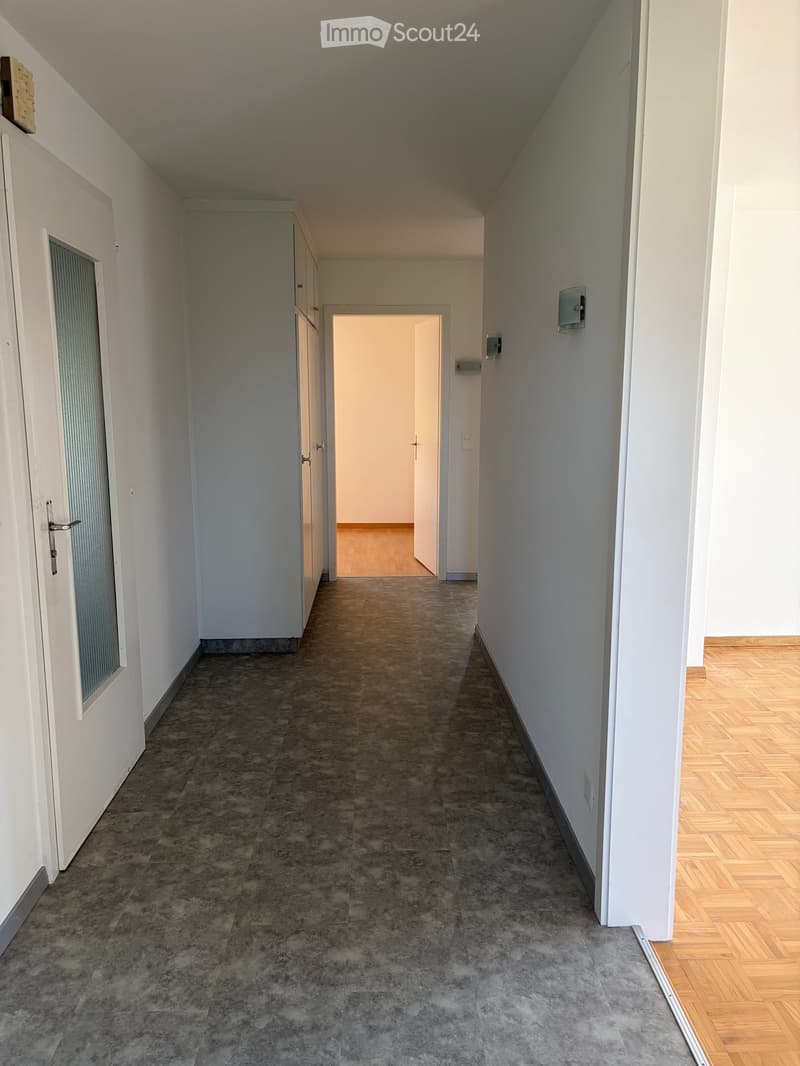 Empty hallway with gray tiled floor, white walls, and wooden flooring visible in the adjacent room. Doors and light switches are present.