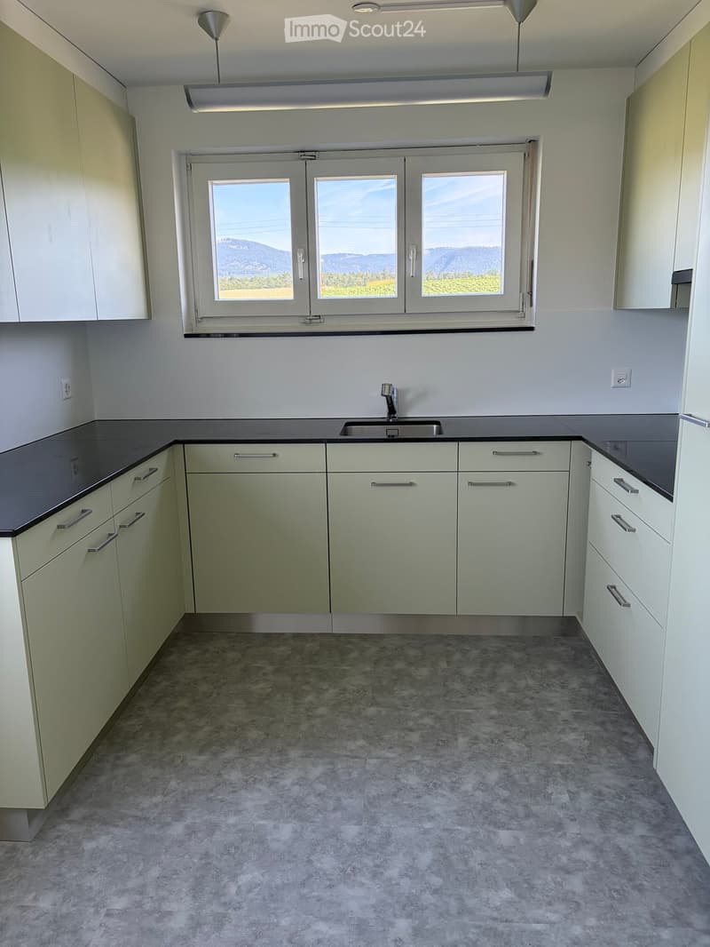 Empty kitchen with light green cabinets, dark countertops, and a sink, featuring large windows with a scenic view.