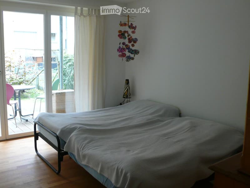 Bright bedroom with wooden flooring, a single bed covered with a light blue duvet, a small table with a pink chair, a wicker basket, and a decorative wall hanging. Large sliding glass doors lead to a garden area.