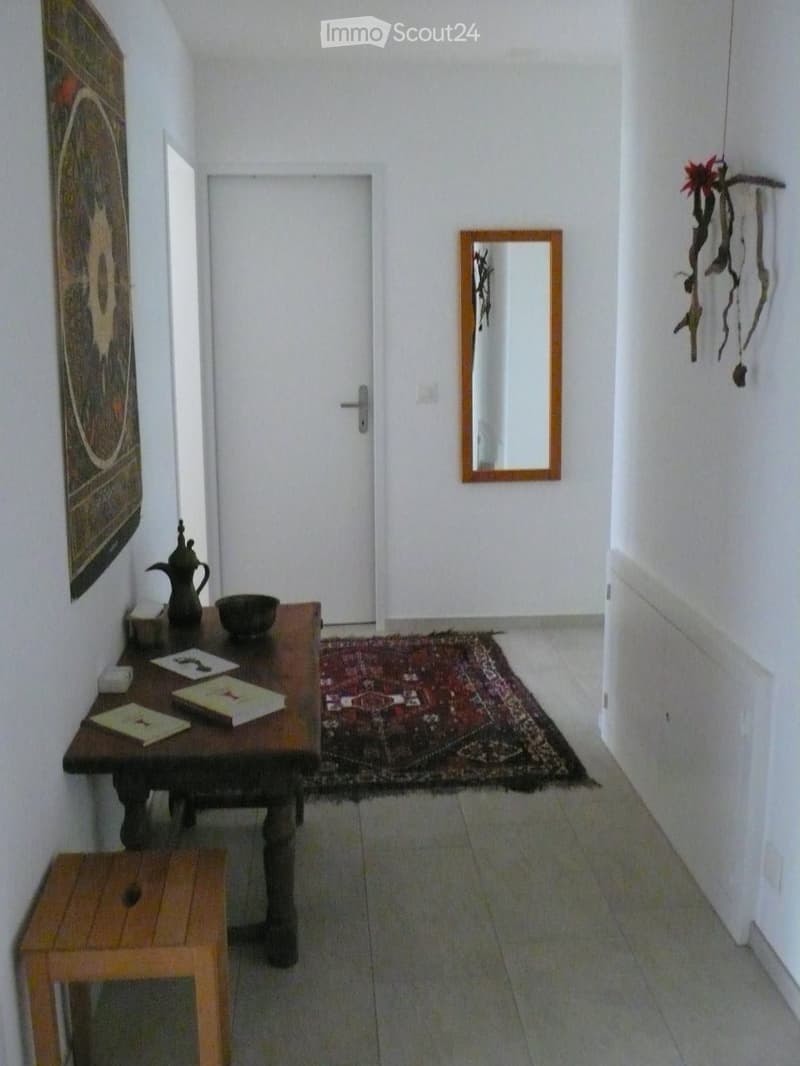 A narrow hallway with a small wooden table holding a vase, bowl, and books, a wooden stool, a mirror on the wall, and a colorful rug on the floor.