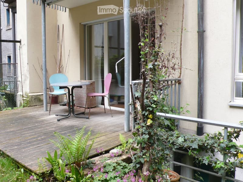 Balcony with wooden decking, small table, two chairs, and a wicker basket. Plants and flowers are visible around the area.