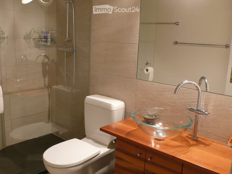 Modern bathroom with a white toilet, wooden vanity, glass sink bowl, chrome faucet, and a glass-enclosed shower area.