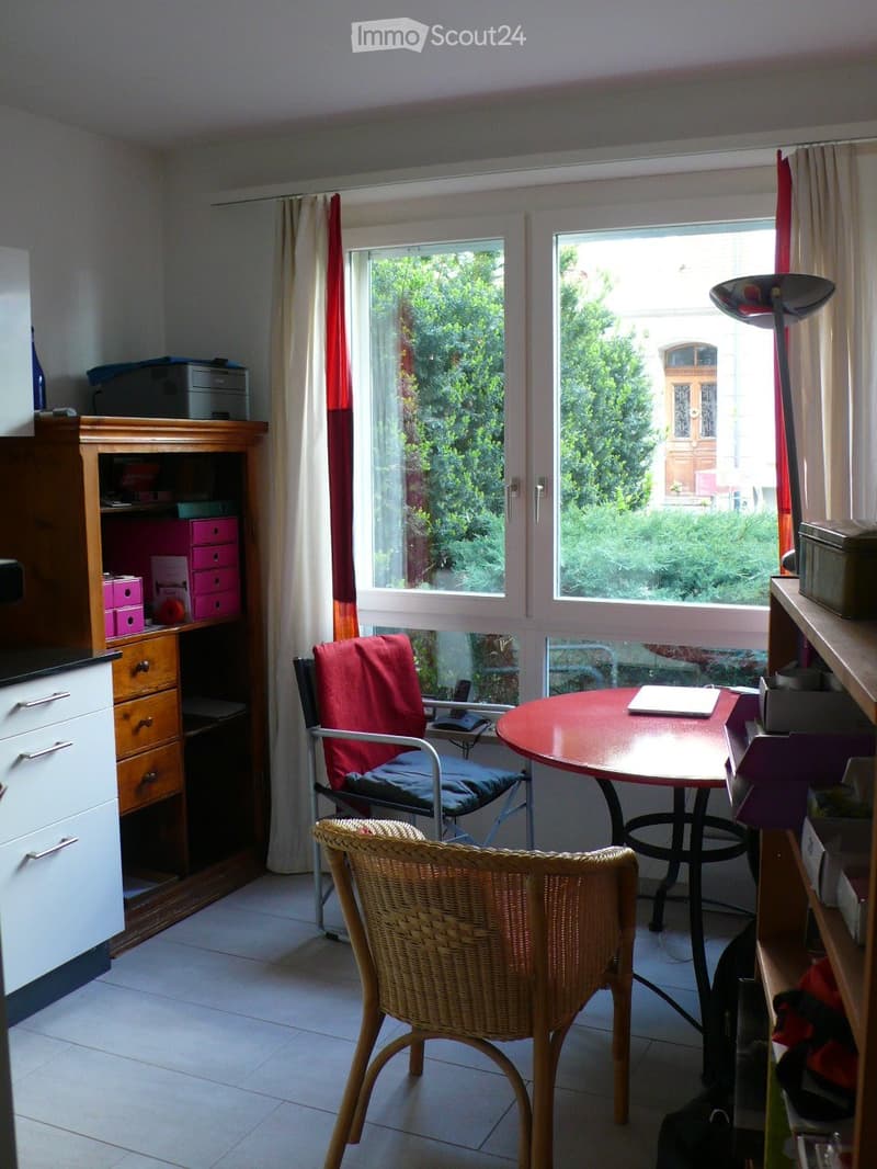 A furnished room with a round table, chairs, a cabinet, and a window with curtains. The room has a tiled floor and a view of greenery outside.