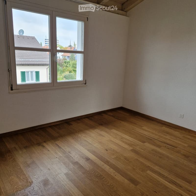 Empty room with wooden flooring and large windows offering a view of neighboring buildings and greenery.